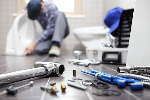 A man performing a plumbing service in the background with plumbing equipment on the floor in the foreground.